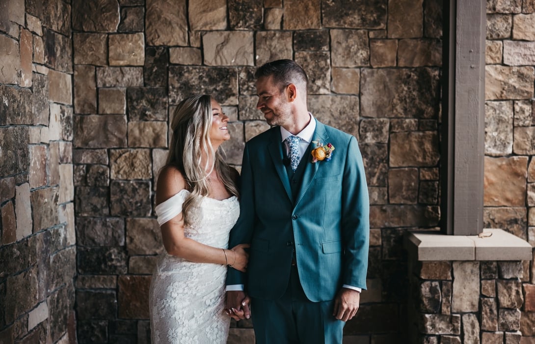 Bride in a white lace dress and groom in a teal suit hold hands and smile at each other against a stone wall backdrop.