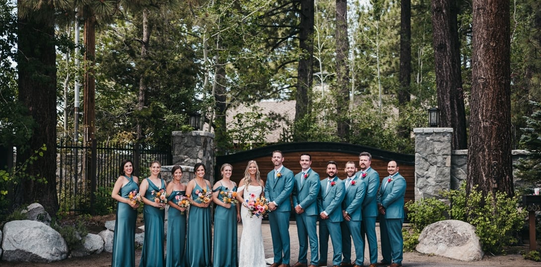 A wedding party stands in front of a stone structure surrounded by trees.