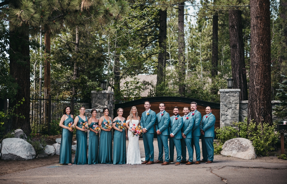 Bride and groom with bridal party in teal dresses and suits, posing outdoors in a wooded setting with bouquets.