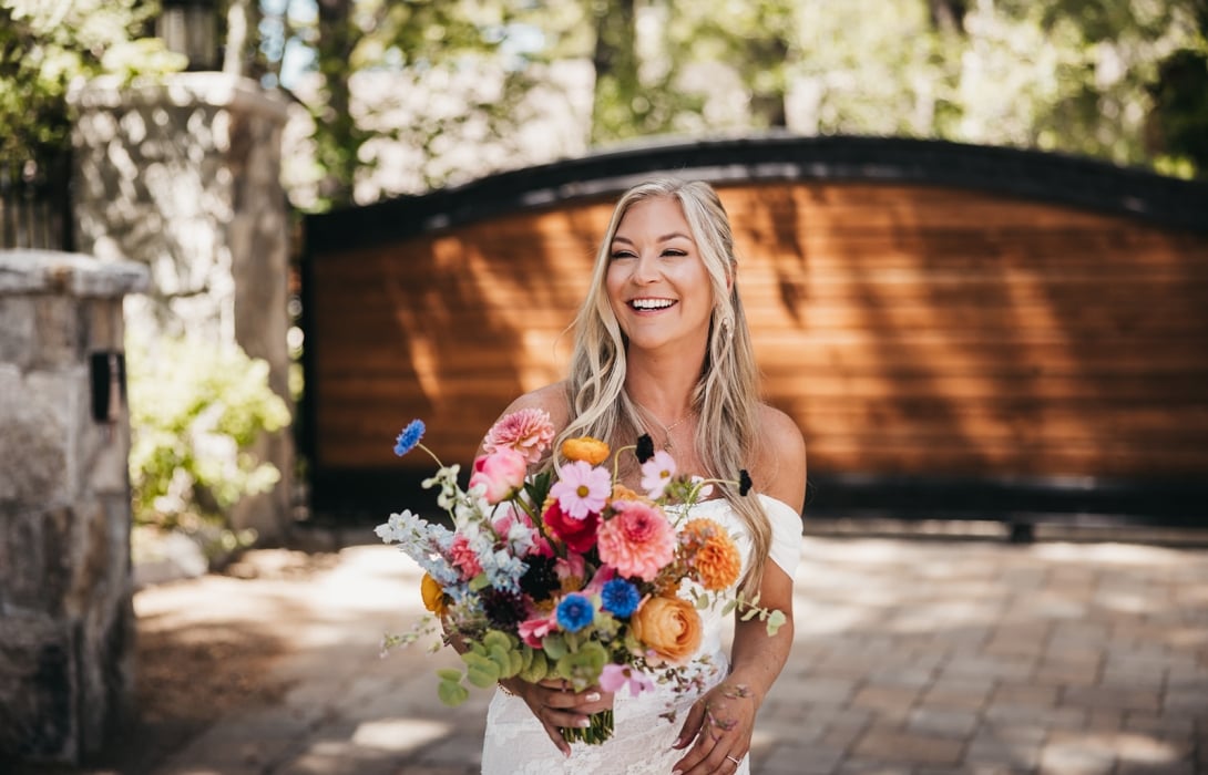 Smiling bride in a white lace dress holding a colorful bouquet outdoors.