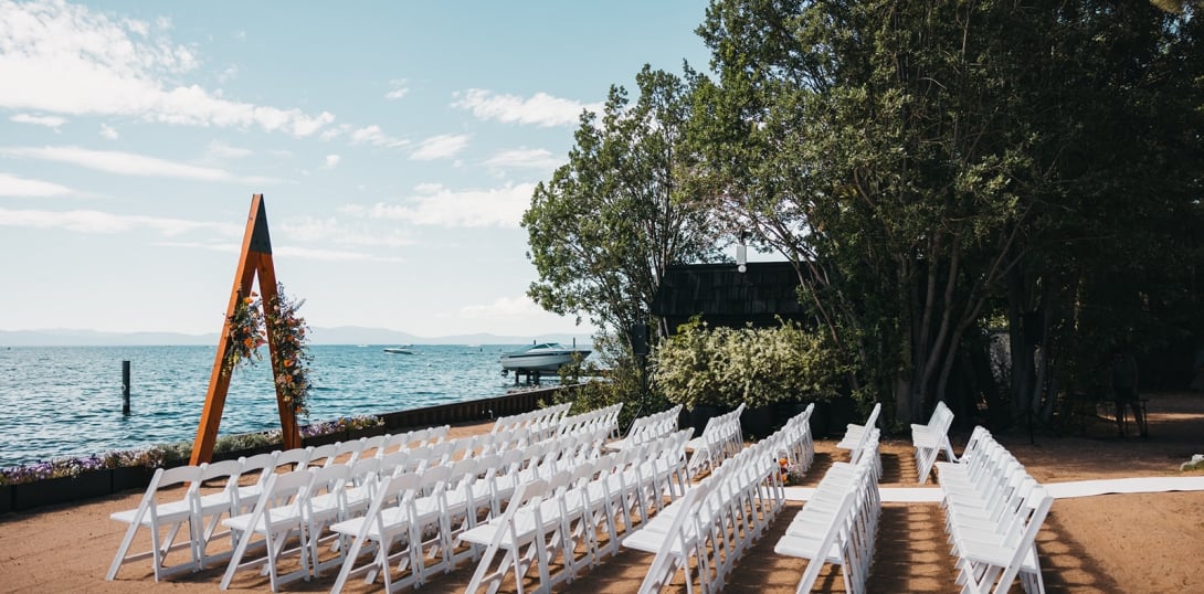 A lakeside wedding setup with white chairs and a floral arch.
