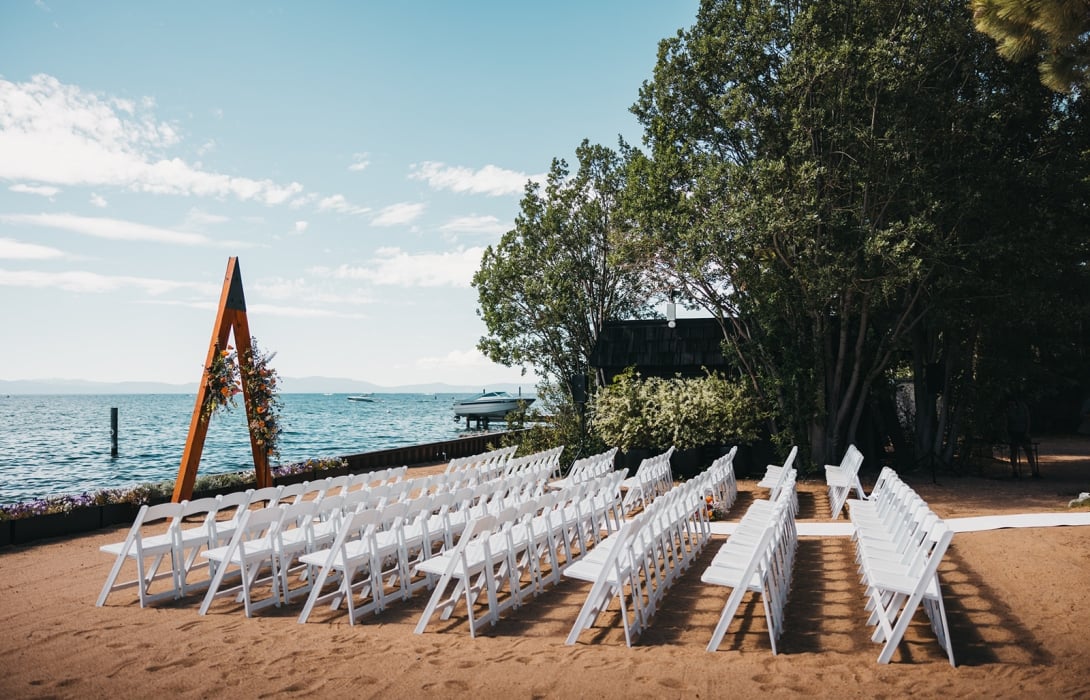 Beach wedding setup with white folding chairs in rows facing a wooden floral arch by the water