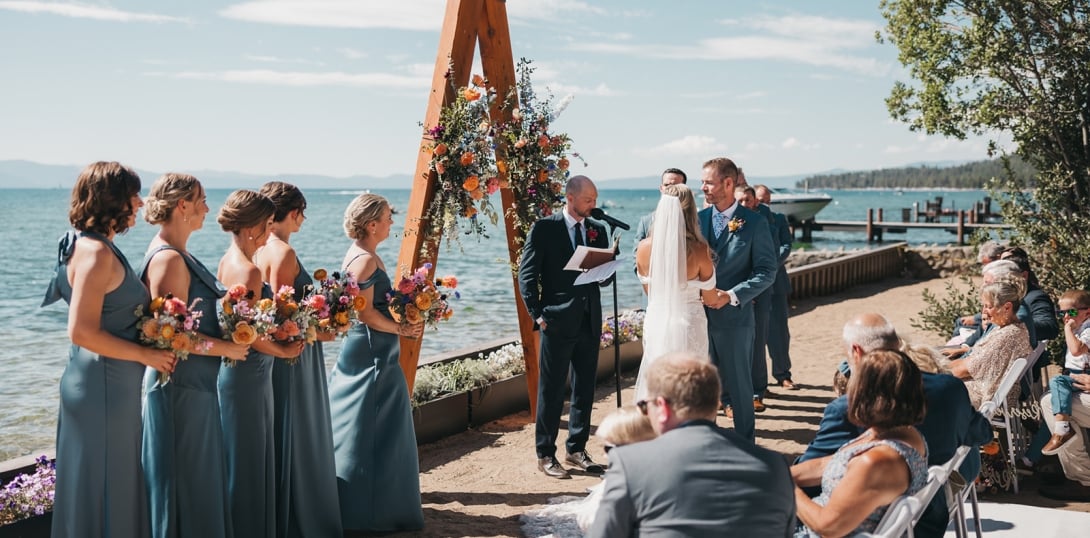 A wedding ceremony taking place outdoors on a beach with a lake and mountains in the background.