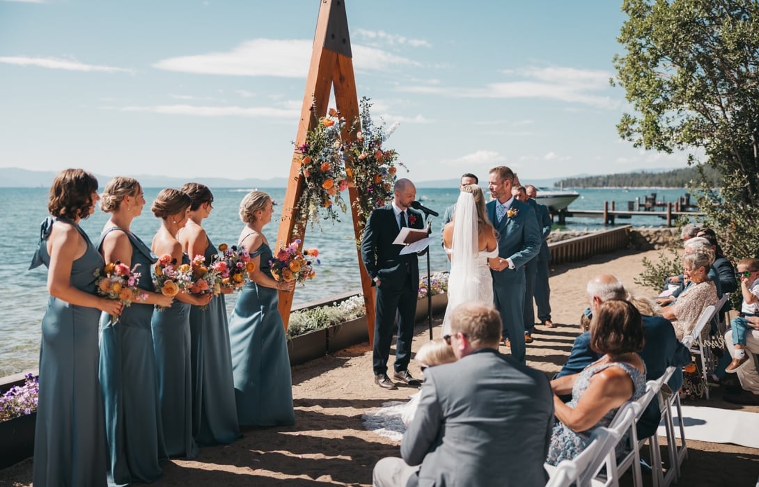 Bride and groom exchange vows by the water under a wooden floral arch; bridesmaids in blue gowns stand to the left holding bouquets while guests look on on the beach.