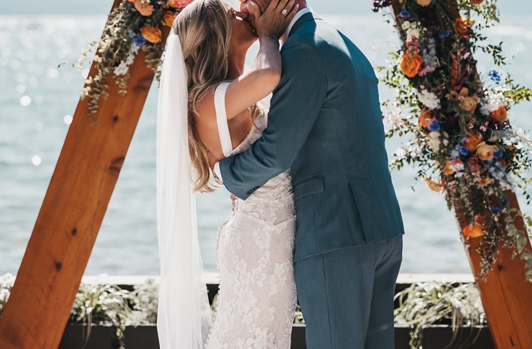 Bride and groom kiss beneath a floral wooden arch by the water, during a lakeside wedding ceremony.