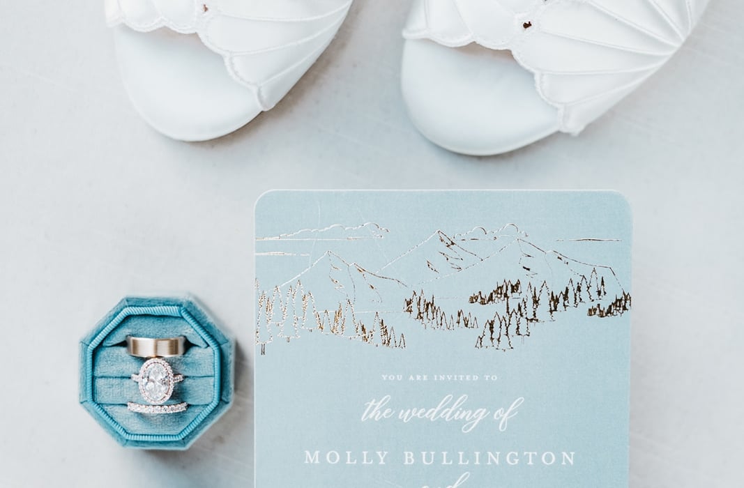 Light blue wedding invitation card with mountain illustration, flanked by white scalloped bridal shoes and blue ring boxes containing an engagement ring and wedding bands.