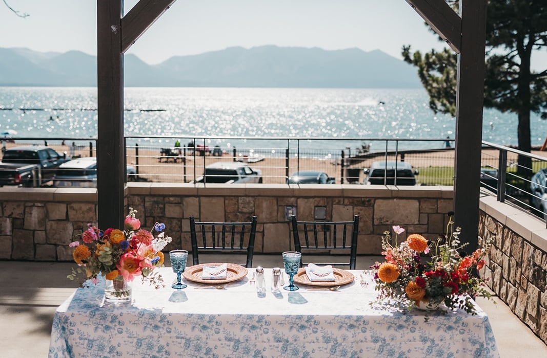 Outdoor waterfront dining table for two with floral centerpieces and place settings, under a wooden arch overlooking the lake and mountains.