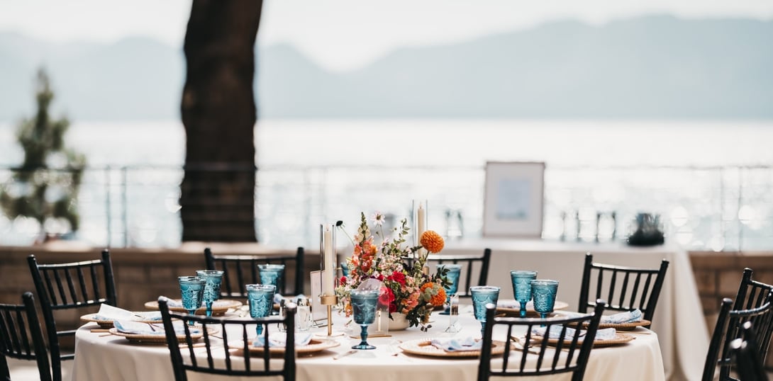 A beautifully set outdoor dining table with a white tablecloth, blue glasses, and a flower arrangement, overlooking a lake and mountains.