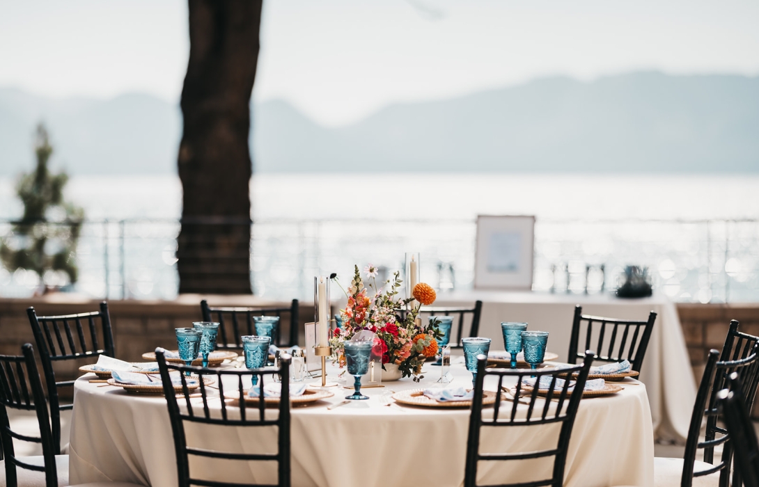 Elegant outdoor dining table by the water with a colorful flower centerpiece and blue glassware