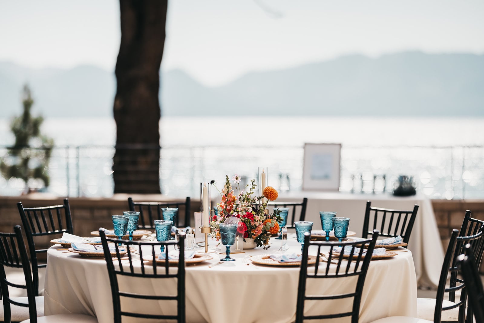 A beautifully set outdoor dining table with a white tablecloth, blue glasses, and a flower arrangement, overlooking a lake and mountains.