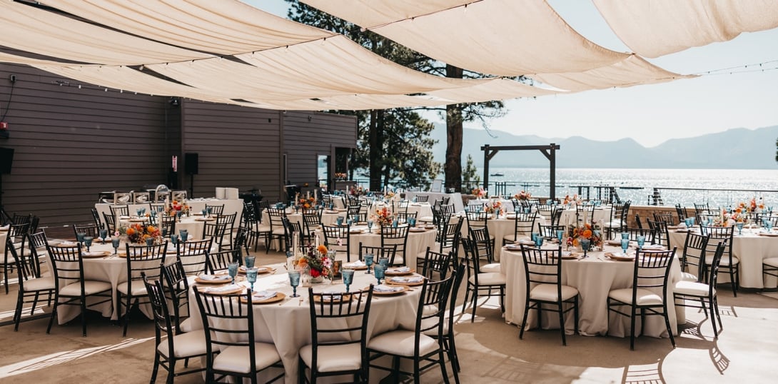 An outdoor event space with white tables, black chairs, and floral arrangements, overlooking a lake and mountains.