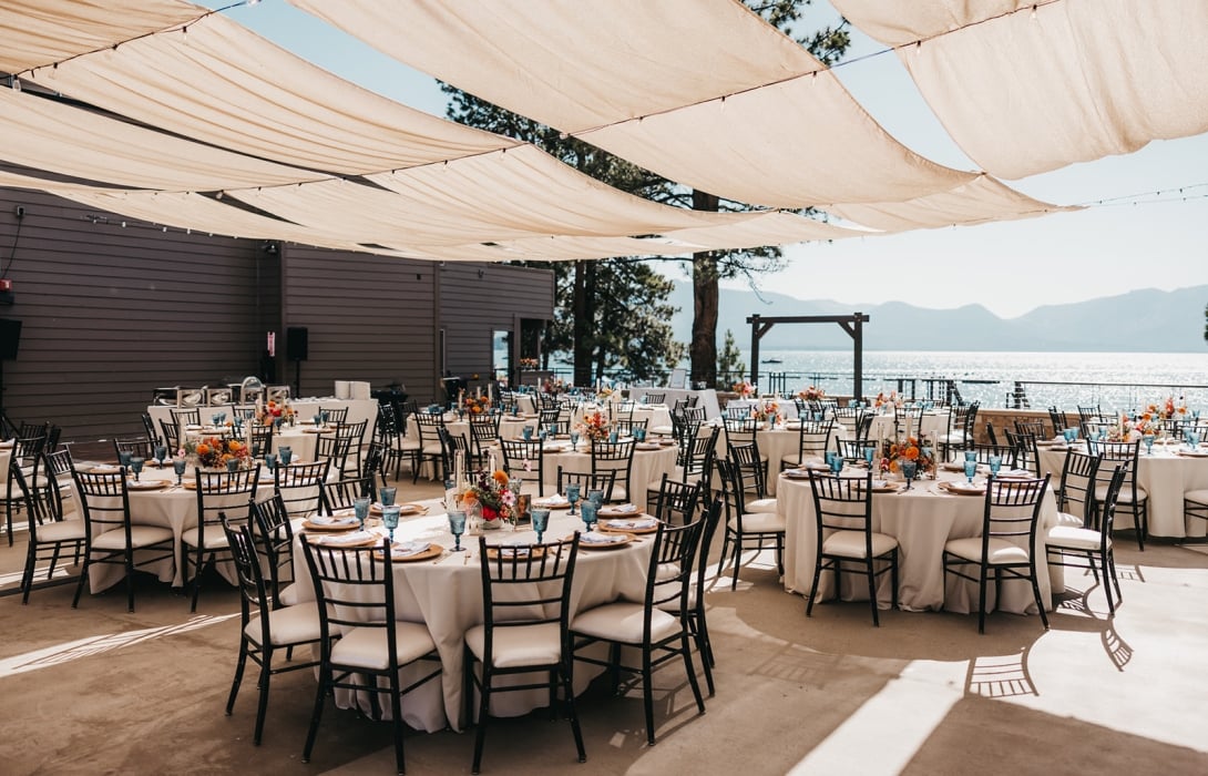 Beachfront outdoor banquet setup with round tables, white cloths, and black chairs under fabric canopies, overlooking the water.