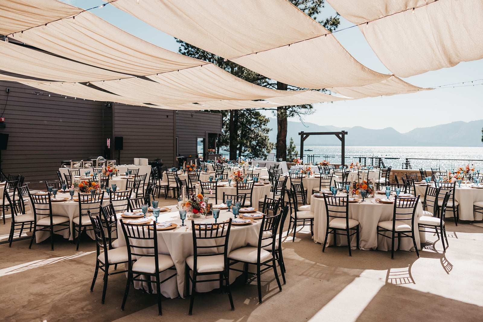 An outdoor event space with white tables, black chairs, and floral arrangements, overlooking a lake and mountains.
