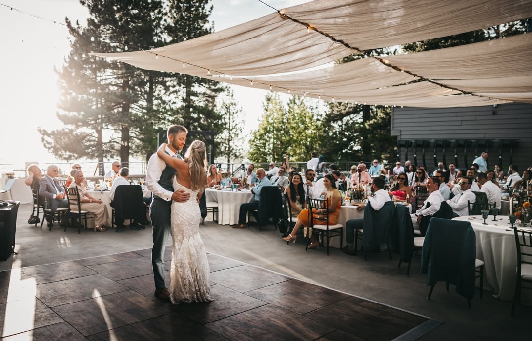 Bride and groom dance together on a wooden dance floor as guests sit at round tables beneath string lights at an outdoor wedding reception.
