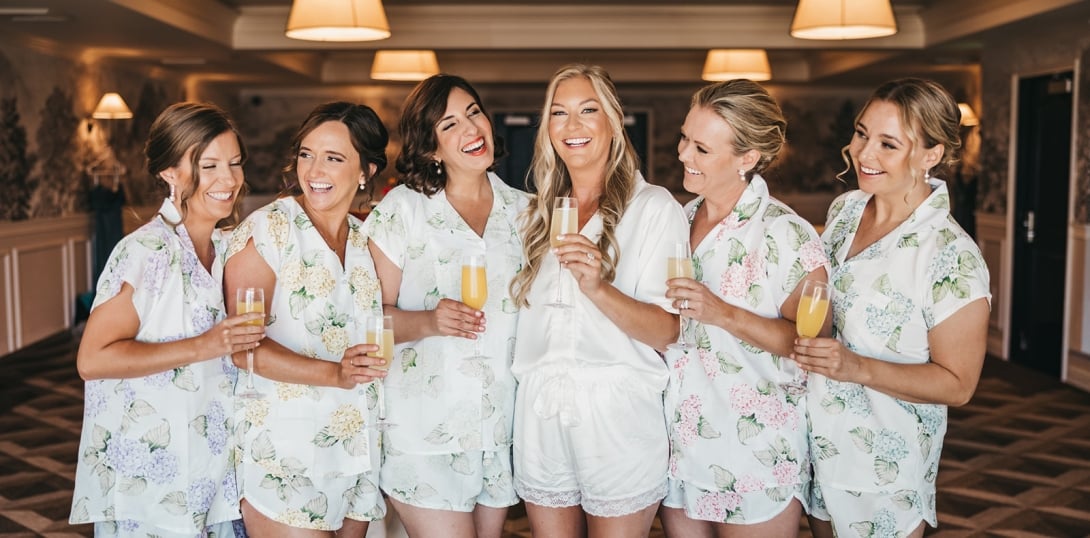 Six women in floral pajamas hold champagne glasses and smile in a hotel lobby.