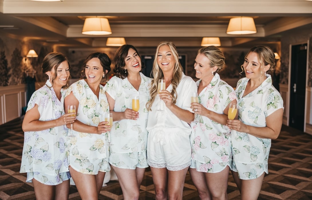 Bride in a white robe surrounded by bridesmaids in floral pajamas, toasting with champagne in a warmly lit room.