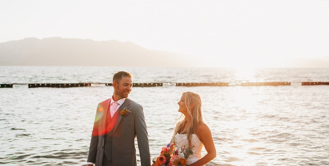 a man and woman in wedding attire standing by the water with mountains in the background