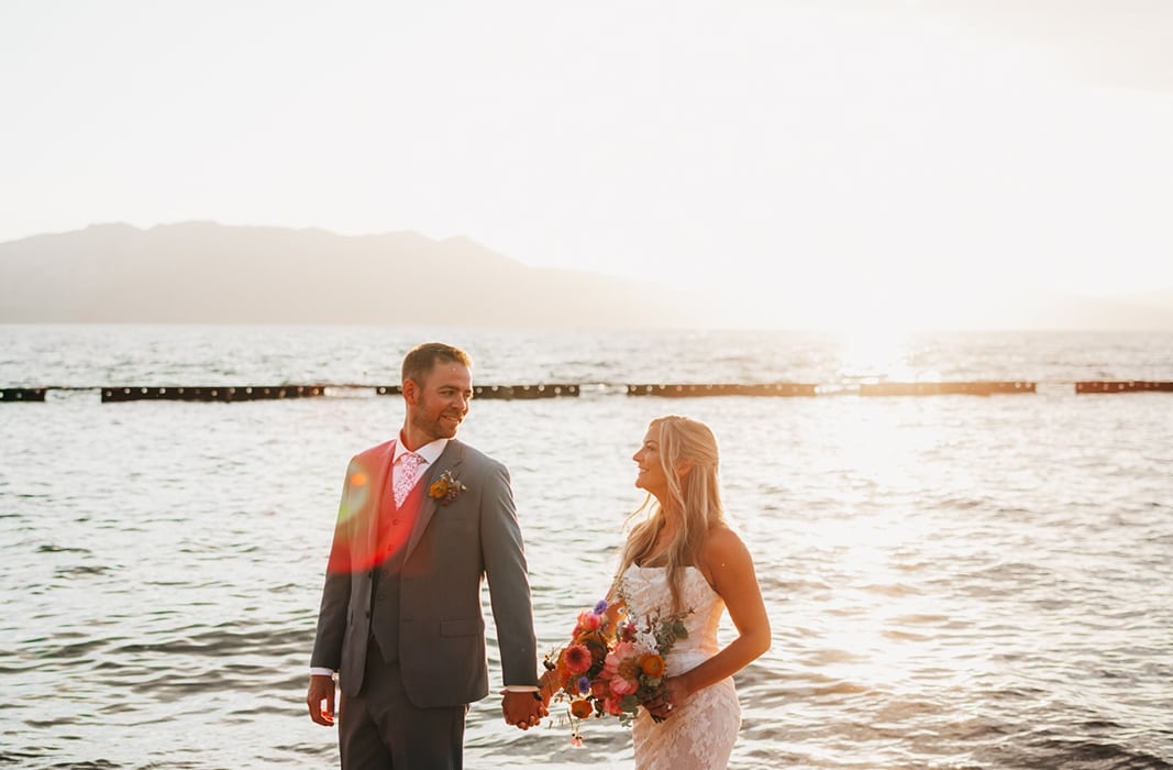 Bride in a lace gown and groom in a gray suit walk hand in hand along a sunset beach, bouquet in hand. (informative)