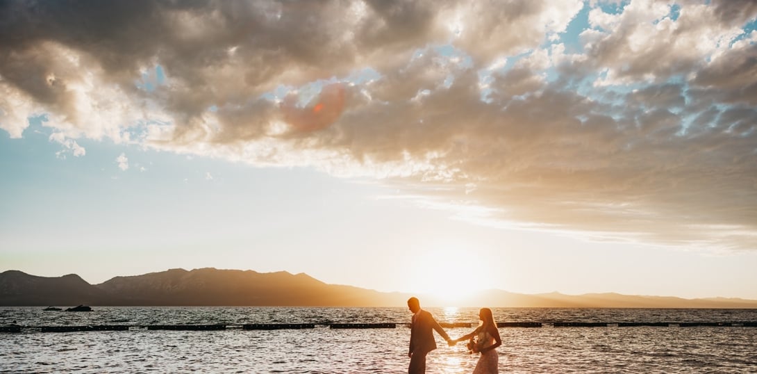 A couple holds hands in the shallow water of a lake at sunset, with mountains in the background.
