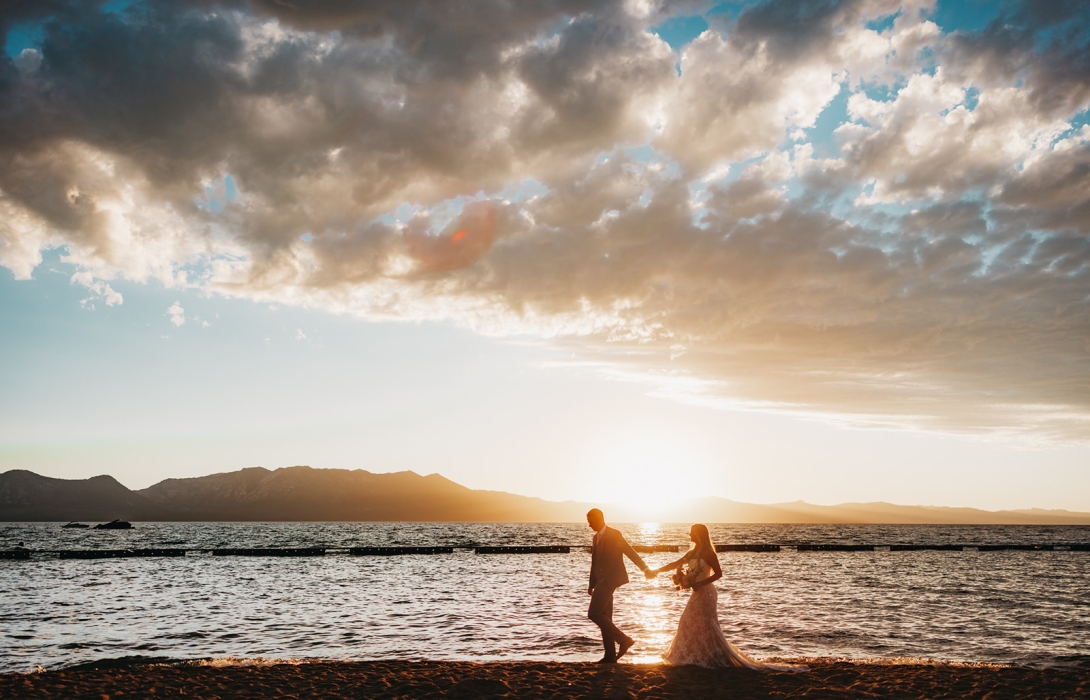 A couple in wedding attire walking hand in hand along the beach at sunset, silhouetted against the water and mountains beyond.