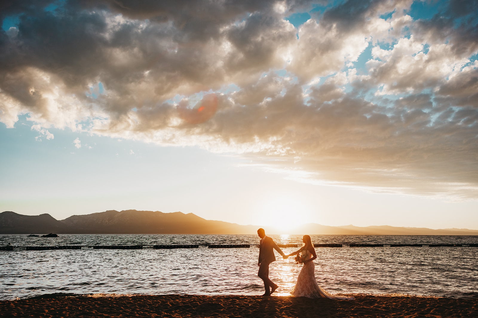 A couple holds hands in the shallow water of a lake at sunset, with mountains in the background.