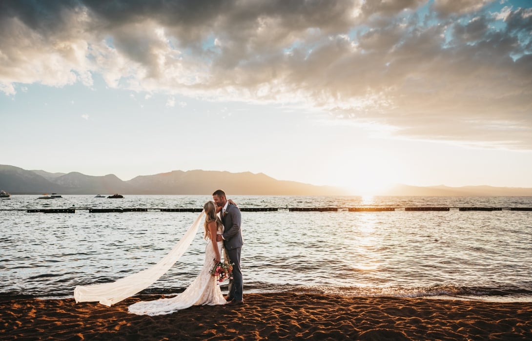 Bride and groom kiss on a sandy beach at sunset, veil trailing behind and bouquet in hand.