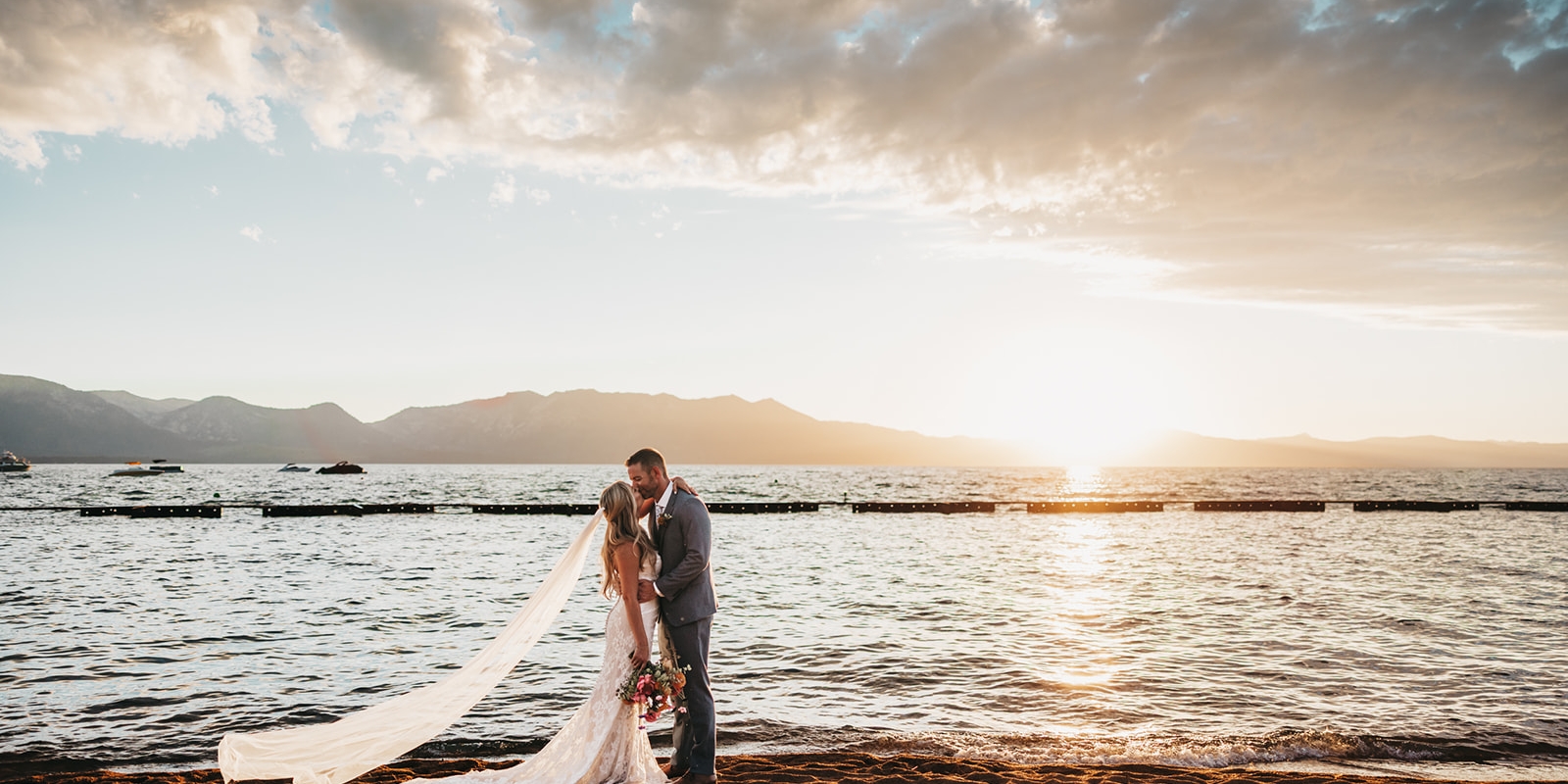 Bride and groom kiss on a sandy beach at sunset, veil trailing behind and bouquet in hand.
