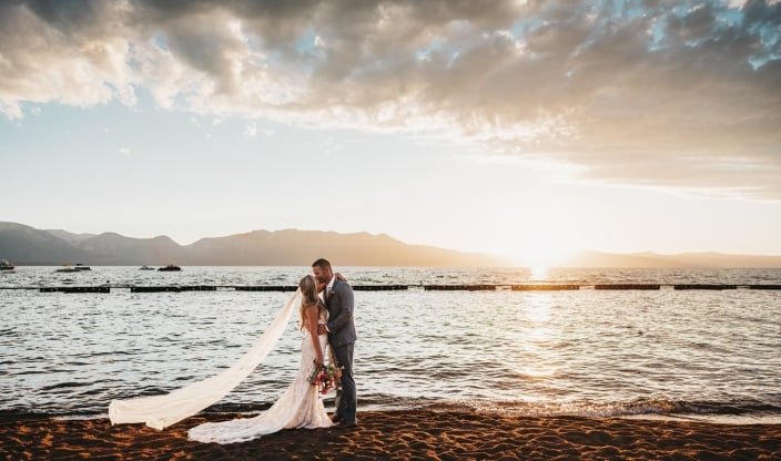 Bride and groom kiss on a sandy beach at sunset, veil trailing behind and bouquet in hand.